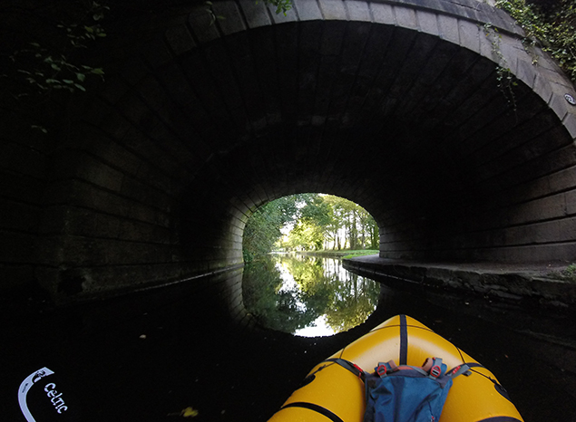 Front of packraft on canal about to go under a stone bridge.